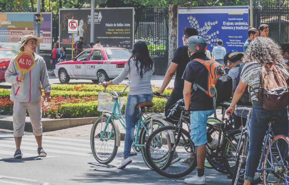 Grupo de ciclistas esperando para cruzar en paso peatonal mientras un auxiliar vial sostiene una paleta de “ALTO” en una avenida de Ciudad de México, promoviendo la seguridad vial y el respeto al ciclista en entornos urbanos.