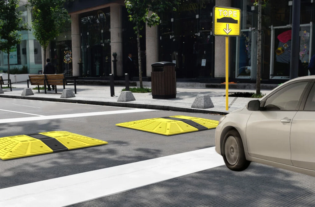 Auto reduce la velocidad frente a señalamiento vial amarillo y tope modular en zona urbana, con señal de advertencia visible y cruces peatonales marcados.